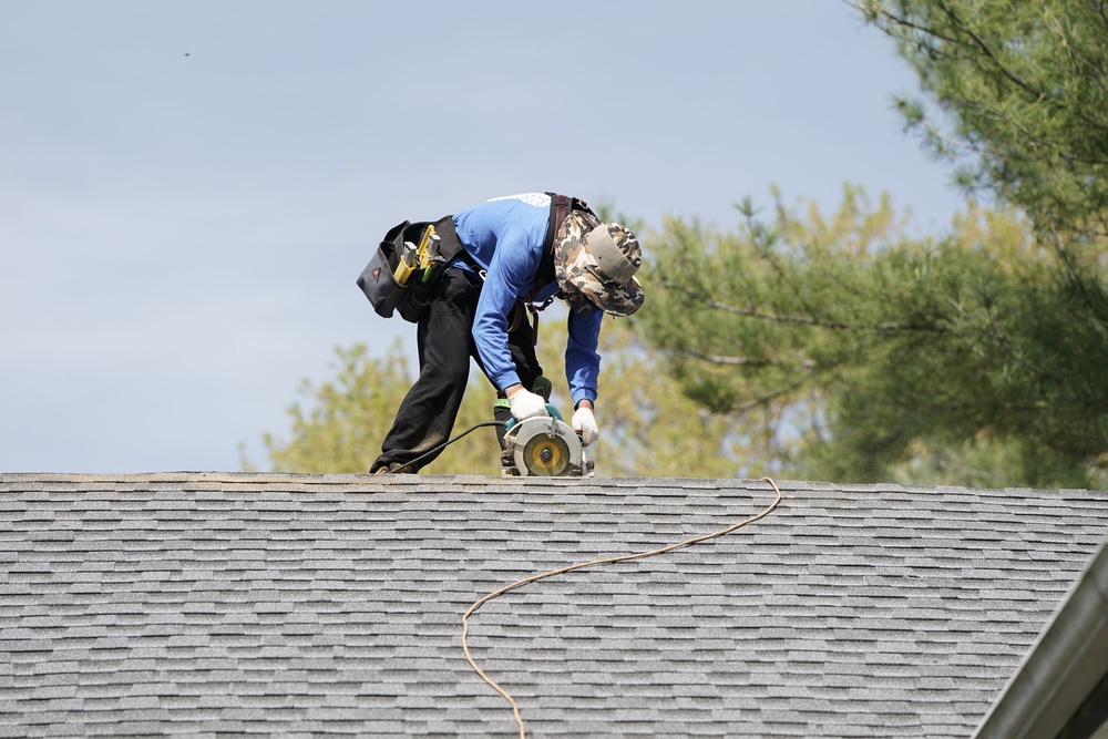 A person in work clothes and a hat uses a circular saw on the roof of a shingled building, with an extension cord trailing behind them.